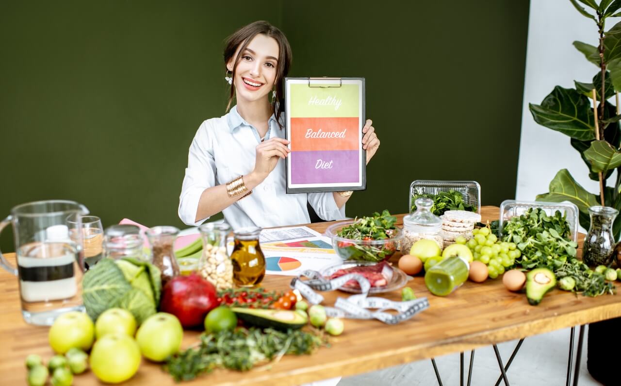 A woman smiling, holding a chart while surrounded by healthy foods ranging from oils, vegetables, meat, and fruits.