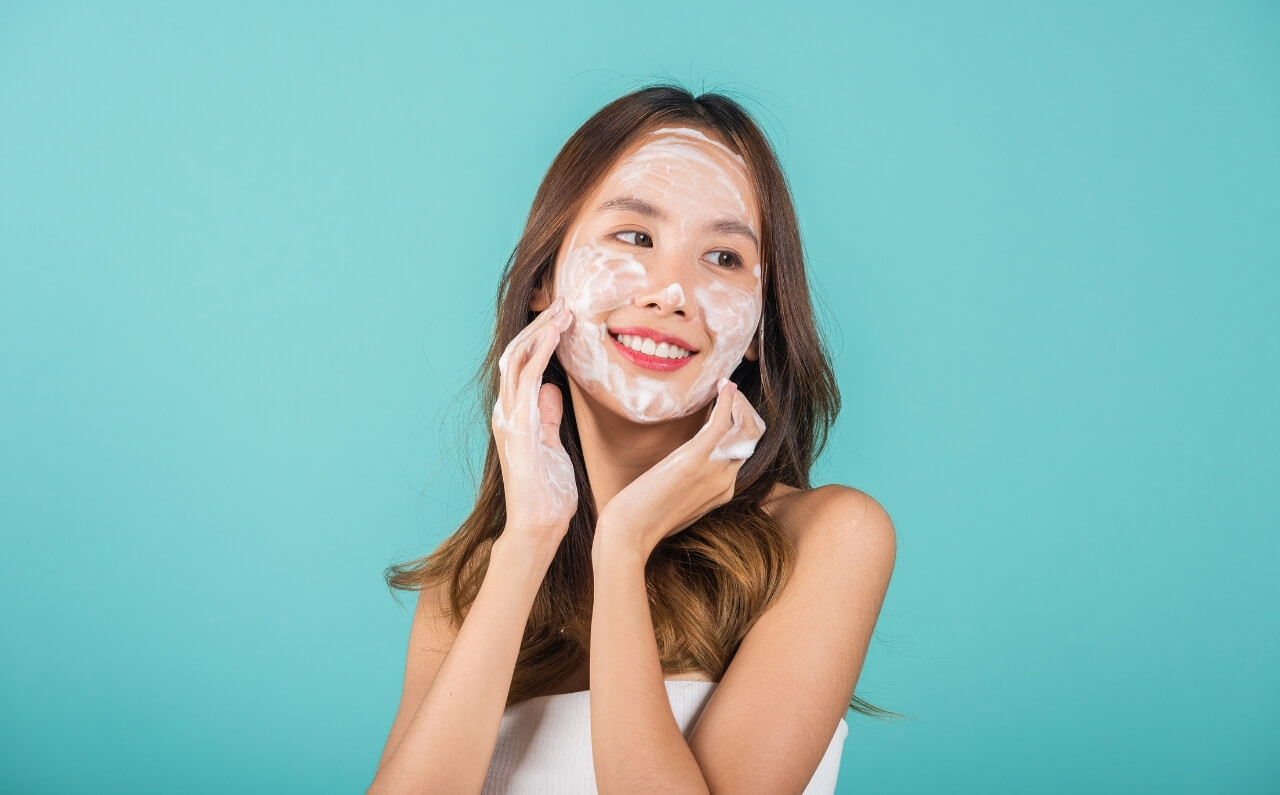 A woman smiling as she cleanses her face.